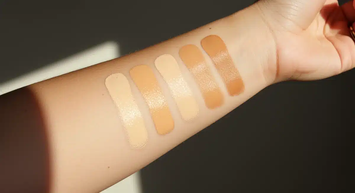 Hand swatching various foundation shades on wrist and jawline in natural light.